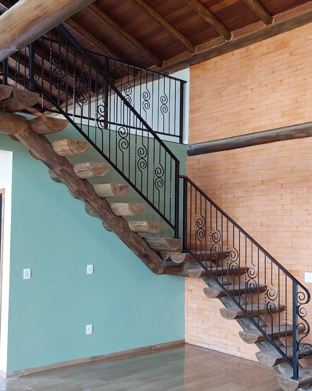Escada com corrimão de ferro preto feito pela Pépe Serralheria, em casa com teto de madeira e parede de tijolinho