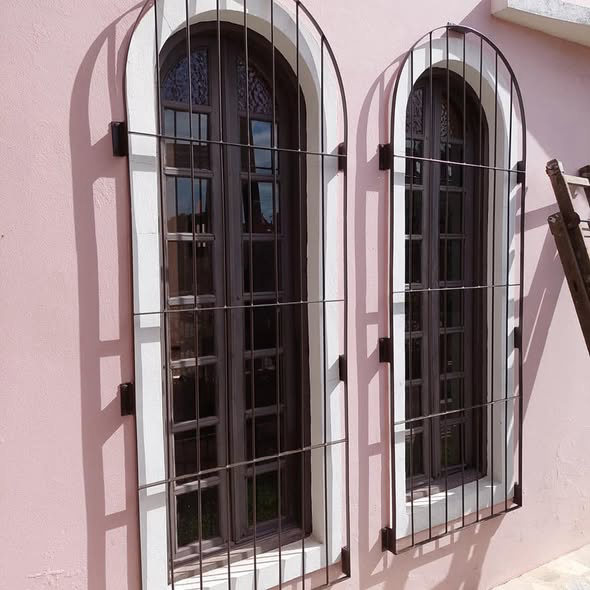 Duas janelas altas em arco com grades marrons numa parede rosa