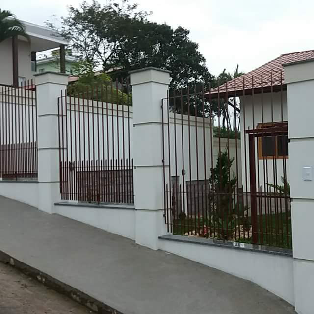 Muro branco com grades pretas entre os pilares, em frente a uma casa
